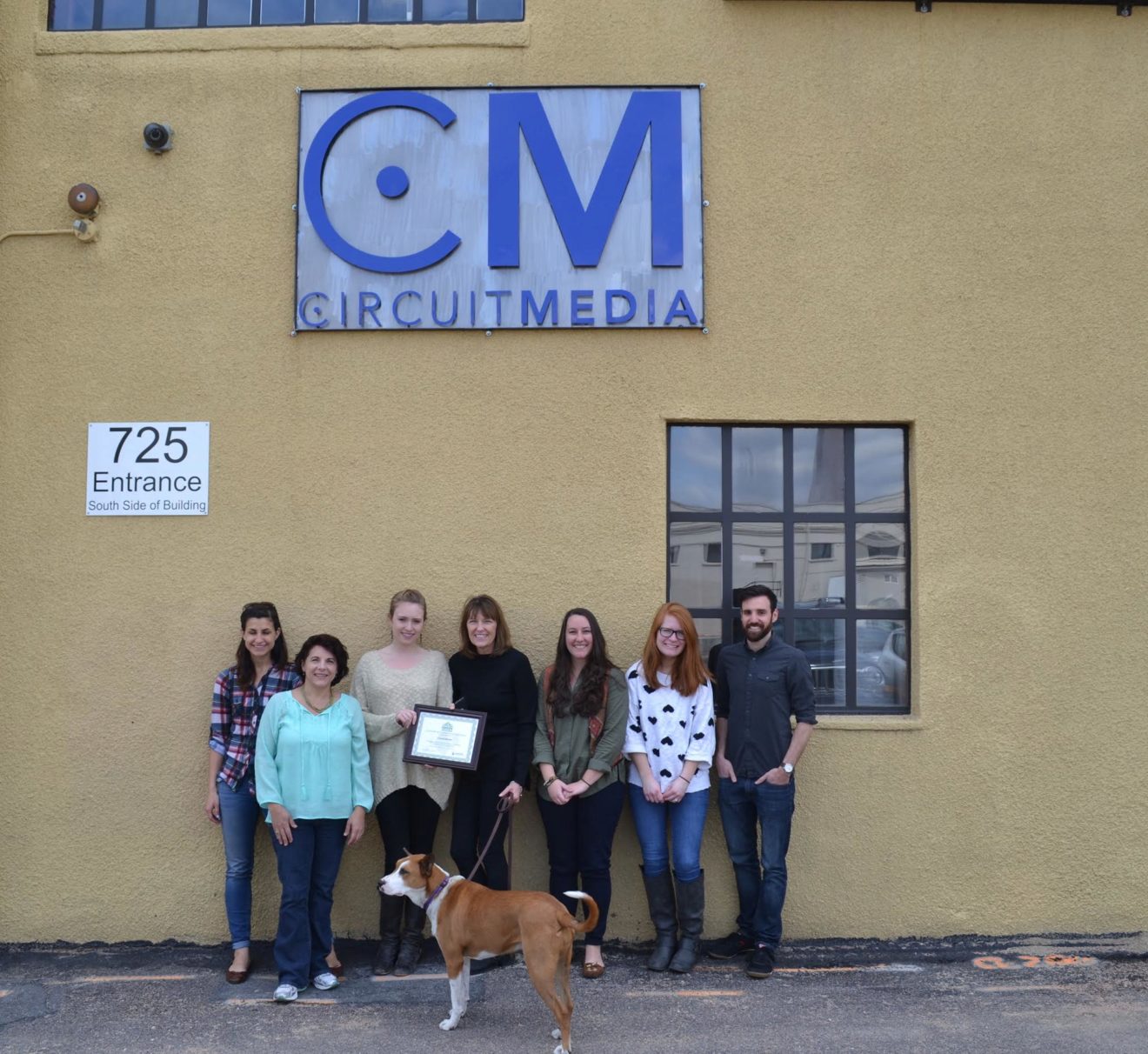 The Circuit Media staff and Paisley pose in front of the Circuit Media sign, holding up their newly earned Green Business Certificate.