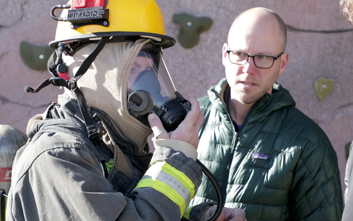 a man in a fire fighter uniform putting his hand to his mask while another man checks his gear to make sure everything is working correctly